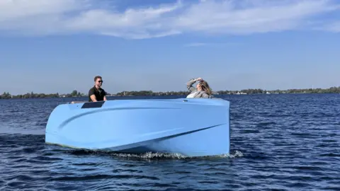 Raw Idea A blue speed boat on a lake with a man in sunglasses at the wheel and a woman at the front.
