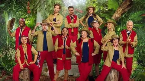 ITV Twelve people can be seen wearing various outfits of red and khaki green, with some wearing large khaki hats. They are posed in front of a jungle backdrop as part of the announcement for the new cast of I'm a Celebrity...Get Me Out of Here!