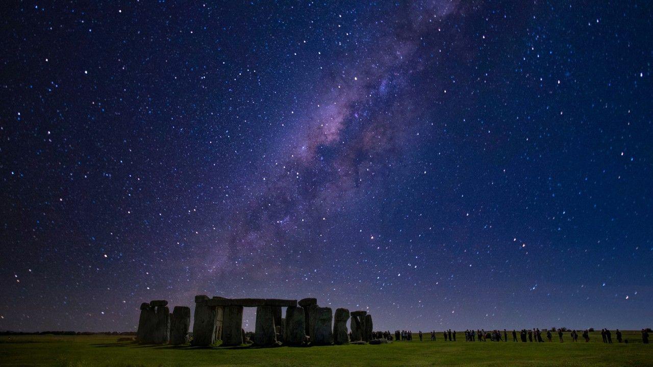 Stonehenge at night on grass, with a line of people queuing under a dark blue and purple starry sky on winter solstice