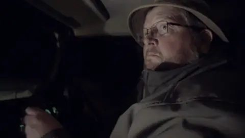 A side view of farmer Morgan Barrett driving in the dark - his face is lit up and he is wearing a thick coat and hat.
