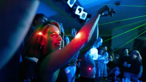 3flavourscornetto A blonde-haired woman smiles and raises her arms as she dances alongside other people. The dark room is illuminated by green and red lasers.