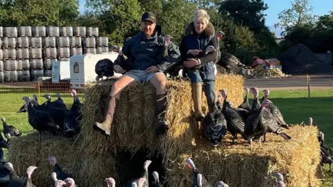 Susan Gorst Susan, a woman in her 50s or 60s, and her son Freddie, probably 20s or 30s. sitting on hay bales. Susan is holding a live turkey and Freddie has his arms wrapped around two more turkeys and there are about 30 more turkeys on and around the hay bales.