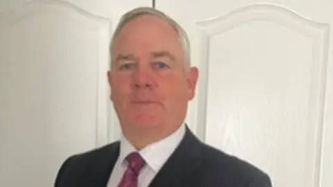 ICLVR A man in a dark suit, white shirt and red tie is stood in front of a white wall. He has grey hair and blue eyes and looks directly at the camera.
