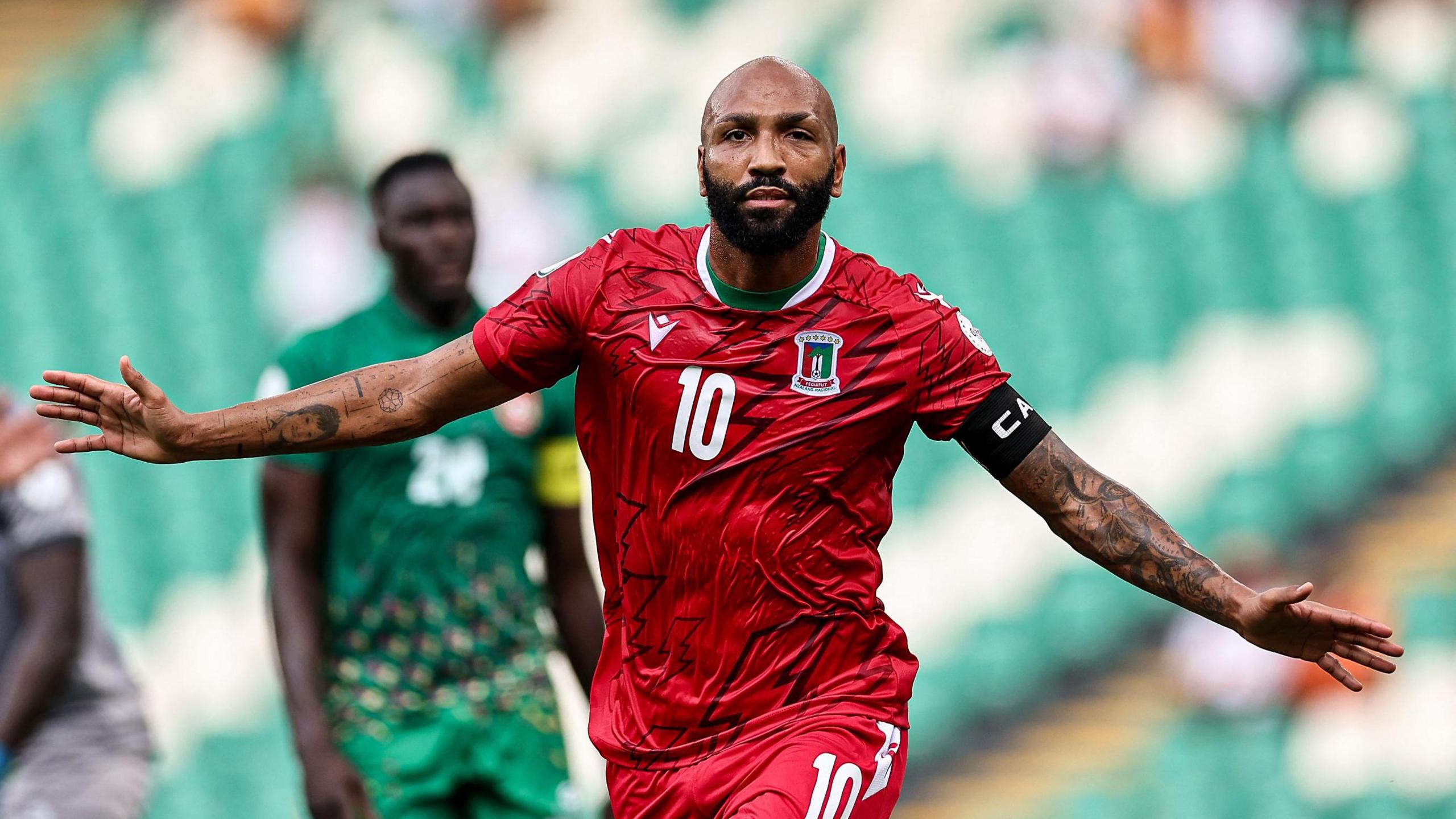 Emilio Nsue, wearing a red Equatorial Guinea shirt with a 10 on the chest, stretches his heavily-tattooed arms out and runs towards the camera in celebration. Nsue has a bald head and thick black beard. A disconsolate Guinea-Bissau player in all-green kit appears blurred in the background along with an empty stand filled with green and white seats