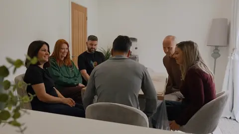 The Southport families seated around a dining table. A grey lamp stands in the corner, and a green potted plant is visible in the foreground on a white counter.