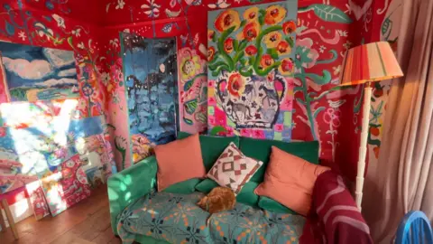 The picture shows a dark emerald green sofa with a ginger cat on it. The walls of the room are red with flowers painted on them. A large painting of flowers also leans against the wall.