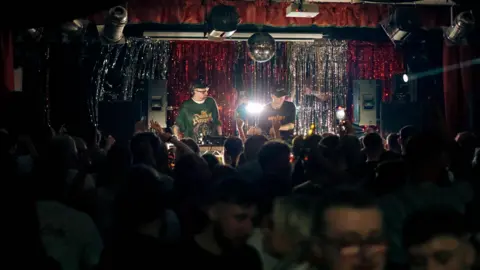 3flavourscornetto Two DJs perform behind the decks on stage. They are both wearing dark jumpers and baseball caps. The dancefloor below them is full. Several speakers are placed to the sides of the stage.