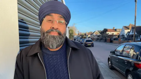 A man wearing a blue jumper, a jacket, a dark blue turban and glasses leans against a wall looking at the camera, with a road and cars in the background