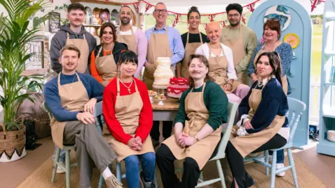 Channel 4/Love Productions The contestants of this year's The Great British Bake Off sitting and standing around a table.