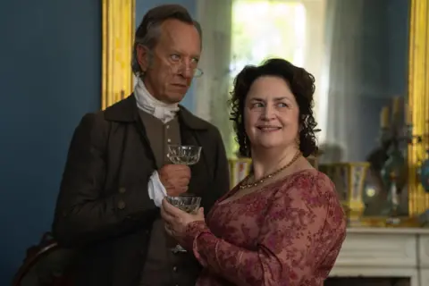 BBC/Bad Wolf/James Pardon Richard E Grant as Mr Bennet, dressed in a suit with a waist coat - he is holding a glass next to Ruth Jones as Mrs Bennet in a floral dress.