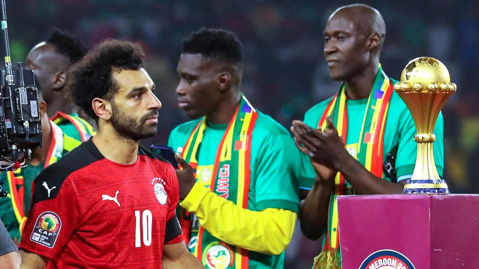 Mohamed Salah visible from waist up wears a dejected expression as he walks past the Africa Cup of Nations trophy, which is sitting on a purple plinth, in his red Egypt kit. Victorious Senegal players and staff wearing mostly green applaud as Salah walks past