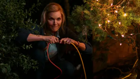 Prime Video Michelle Pfeiffer holding two electric cables for Christmas lights and plugging them together with a mischievous look on her face