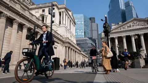 Getty Images A man in a suit rides a bike in through the city in London with the Bank of England building behind him