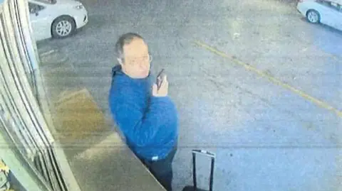 Providence Police/Handout via Reuters A person holding a phone and wearing a blue jumper stands next to a wheeled suitcase in an underground car park with cars in the background.