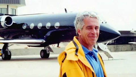 US Department of Justice/PA Jeffrey Epstein, a man with grey hair wearing a bright blue polo shirt and an orange anorak, smiling broadly as he stands in front of his private plane - a black jet with chrome detailing on the wings and around the engines, with five porthole-style windows visible on the right-hand side.
