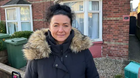 Phil Harrison / BBC A woman with dark hair and a black coat. She is standing in front of a house.