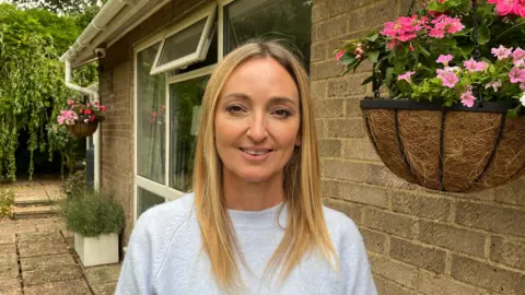 Nicola Haseler/BBC Claudia Pixley, a woman wearing a light blue top with long blonde hair, stands outside her home with a hanging basket next to her.