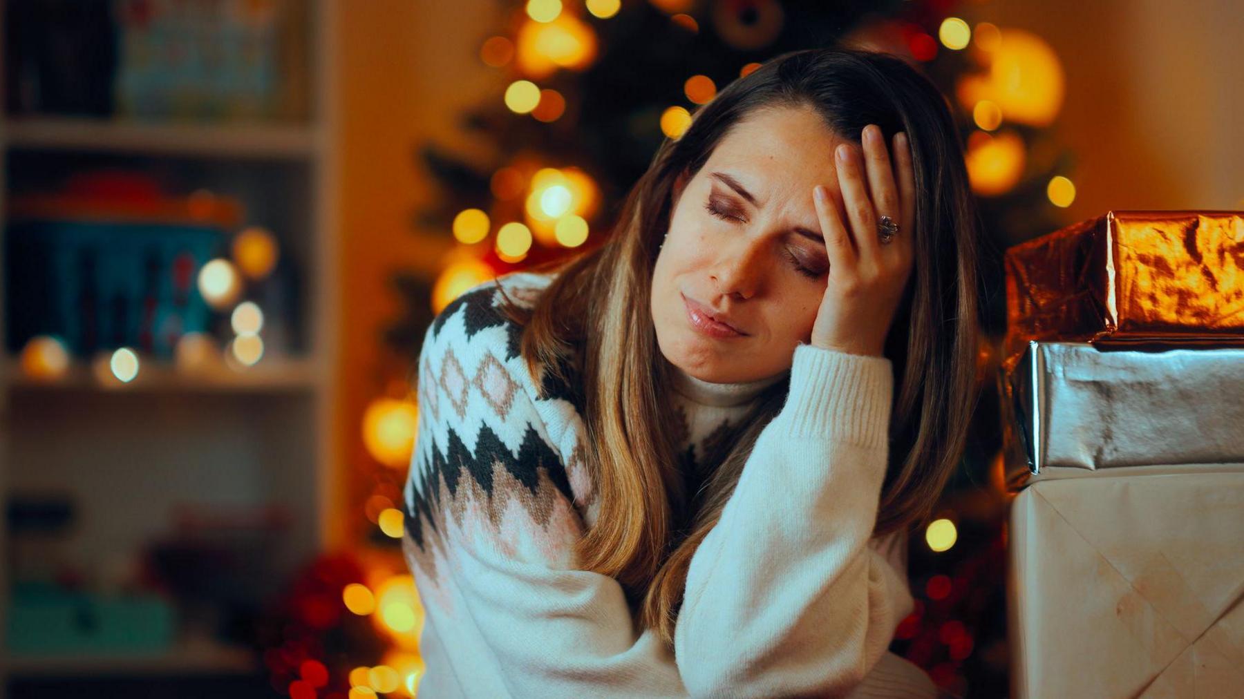 Woman with head in her hands in front of a Christmas tree looking overwhelmed