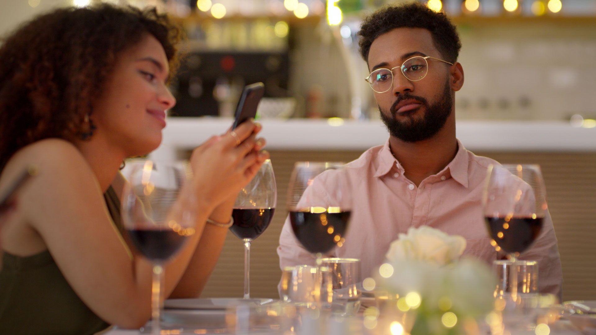 A young couple sit at a restaurant table with glasses of wine whilst the woman is using her mobile phone and the man is looking bored and dejected.