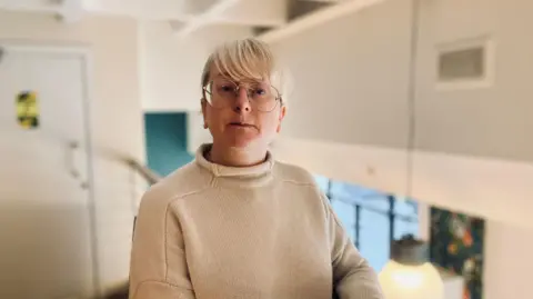 Michelle Langan, who has blonde hair, glasses and is wearing a cream top, is sitting on a chair on the mezzanine level of the cafe she runs. Next to her is a pot plant and a pink, glass-topped table.