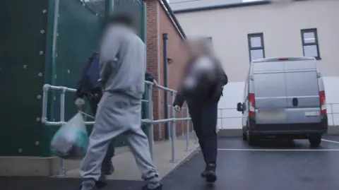 Home Office A man in a grey track suit is escorted into the centre from a van. One of two officers flanking him is holding a plastic carrier bag of belongings.