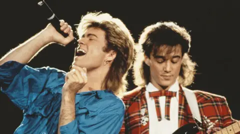 Michael Putland Hulton Archive via Getty Images George Michael (left) wears a blue shirt and sings into a microphone he is holding up to his mouth, while Andrew Ridgeley looks down at a guitar he is playing out-of-frame wearing a tartan suit, as they perform together live on stage in 1985.