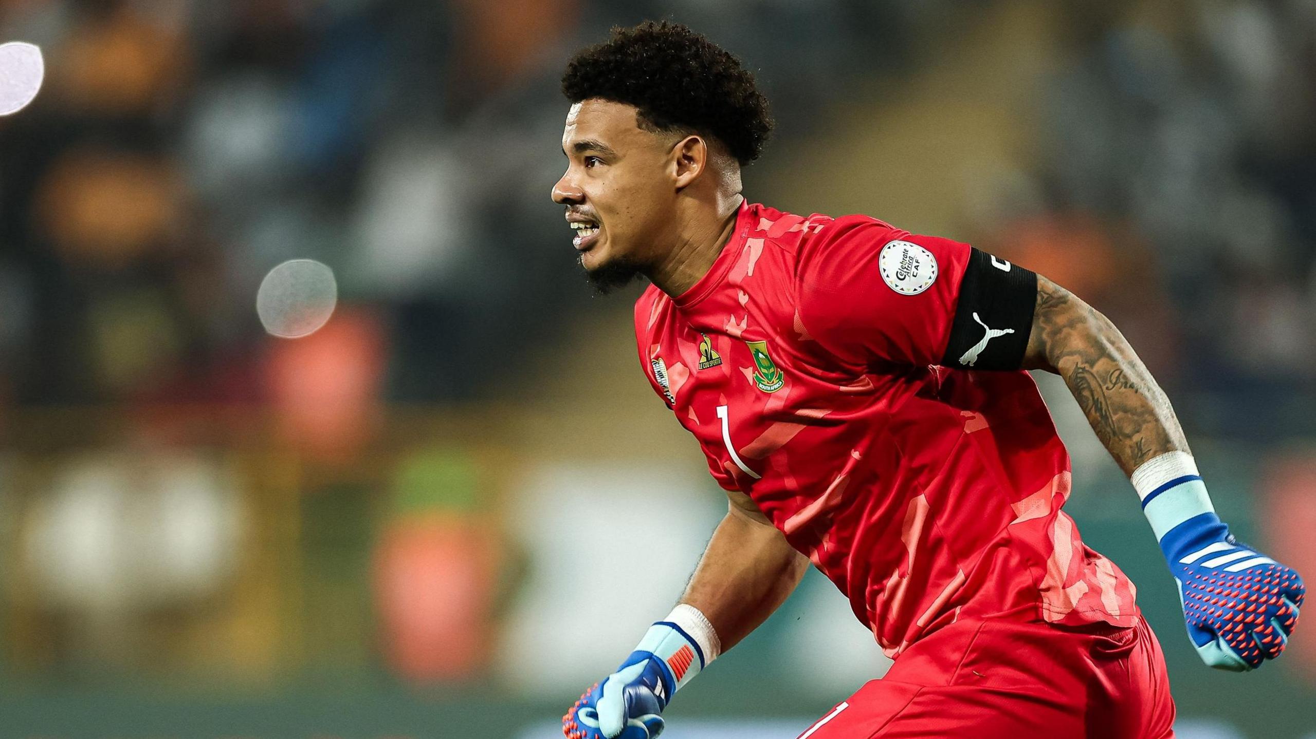 Ronwen Williams, wearing a red South Africa kit and blue goalkeeping gloves, runs towards the left of shot with a slight grin on his face