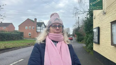 A woman stands on a village street wearing a pink hat and matching pink scarf, along with glasses and a dark coat. Behind her are brick houses, a grassy verge.