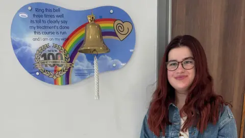 Harriet Trewhitt Harriet, a woman with long, wavy red hair, is standing indoors next to a wall-mounted plaque featuring a large golden bell with a white rope attached. The plaque has a colourful rainbow design and a heart symbol.