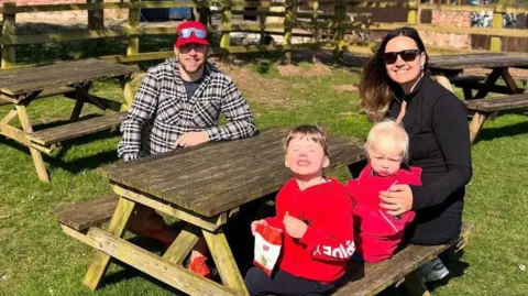 Andrew and Zoe Family photograph of dad Andrew, mum Zo&euml;, and their sons Joey, who is four, and his little brother, two-year-old Tommy. It is a sunny day and they are sitting around a wooden picnic bench