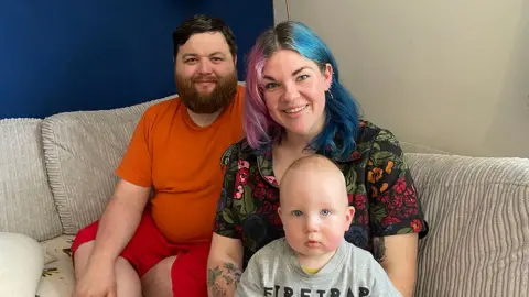 Tom, Laura, and their baby son George, are sitting together on a light-colored sofa in a living room. Tom is wearing an orange T-shirt and red shorts, while Laura has brightly colored blue and purple hair and is wearing a black shirt with a floral pattern. The baby, seated in front, is wearing a light gray shirt with the word “FIRETRAP” printed on it. The background includes a blue wall and a beige wall, creating a contrasting backdrop.