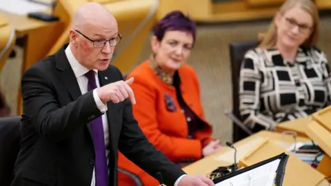 PA Media John Swinney, who is bald with dark-rimmed glasses, speaks at a podium in the Scottish Parliament. He is pointing downwards with his right hand, held out in front of him, with cabinet ministers looking on in the background. He is wearing a dark suit, white shirt and purple.