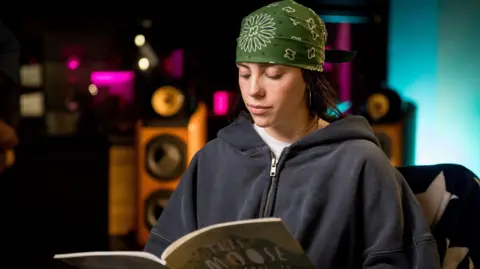 Billie Eilish sits in an armchair and looks down as she reads from a picture book. She wears a baggy blue hoody and a dark green bandana with white shapes forming a pattern on it. In the background recording equipment and speakers can be seen.