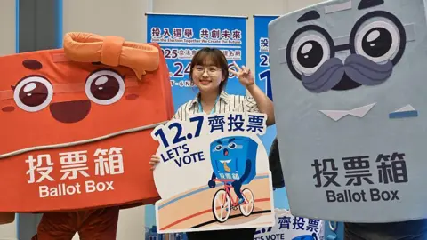 VCG via Getty Images A woman in a striped shirt poses with a sign that says "12.7 Let's vote" with two life-size cartoon mascots of ballot boxes