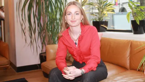 Ivee Blonde-haired Lydia Miller smiles while looking into the camera. She is wearing a bright-red shirt and sits on a leather sofa.
