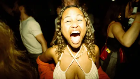 3flavourscornetto A woman opens her mouth widely as she looks directly into the camera as she dances. She has shoulder-length brown hair and is wearing a white top.