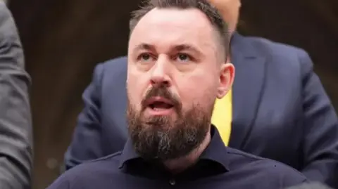 PA Media Martin Hibbert sits speaking. A microphone with a fluffy cover is in the foreground. People in suits stand behind him.