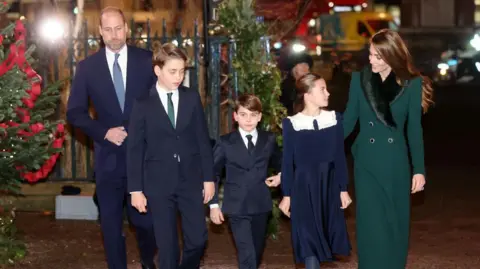 PA Media Prince William, Prince George and Prince Louis are seen walking outside the Abbey wearing suits, shirts and ties, while their sister Charlotte, wearing a dress with a peter pan collar, looks up at their mother Catherine, who wears a green coat with a furry collar