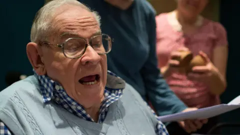 Alamy Baxter reading from a script in the foreground while actors watch from behind. He is seated, wearing a blue jumper