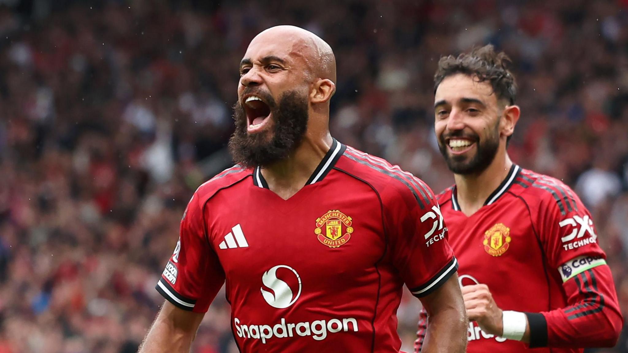 Bryan Mbeumo, with a shaven head, a bushy black beard and wearing a red Manchester United shirt, screams in celebration during a football match. Team-mate Bruno Fernandes is seen smiling running behind him. Both men are viewed from waist up with a blurred stadium crowd in the background