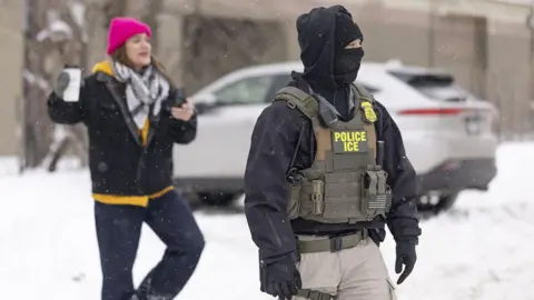 Andaou via Getty Images An ICE agent in a black balaclava and ICE stab vest is standing amid the snow. Out of focus, behind the agent, a woman in a pink hat and keffiyeh, holding a take-away coffee cup, can be seen.