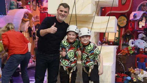 Andres Poveda A man with short brown hair and a grey beard wears black jeans and a black jumper. Two young boys wearing identical white helmets and green and black t-shirts stand to his left.