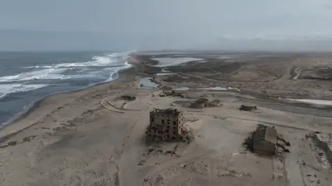 An aerial view shot from a drone of the coast line near Alexander Bay. The shell of a former mine building can be seen in the foreground amid a damaged landscape up against the sea.