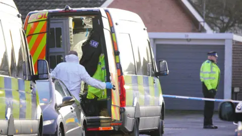 PA A police forensics officer in white overalls is seen from the back, opening a van door. There is another police van parked next to it. In the background there is a police cordon across the road, with a police officer behind it. It is a residential street, with a house and a garage partially visible.
