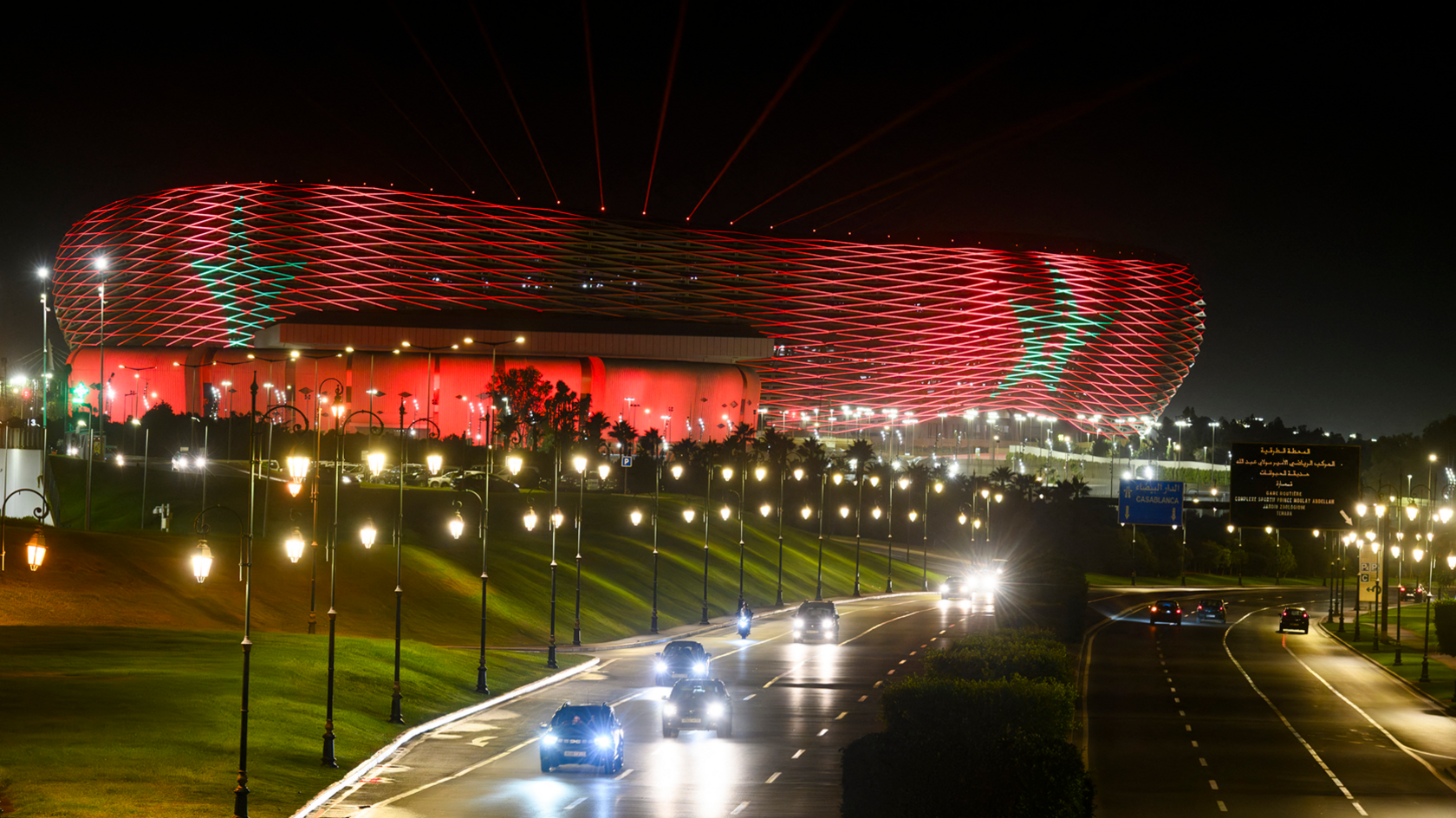 The exterior of the Prince Moulay Abdellah Stadium is lit up at night in the colours of the  Moroccan flag, with a red base colour and two green five-point stars. The stadium towers over a nearby road lined by street lamps, on which several cars are coming towards the camera with their headlights shining