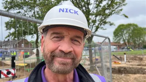 Nick Knowles wearing a hard hat and smiling at the camera. His hat has 'Nick' written on it