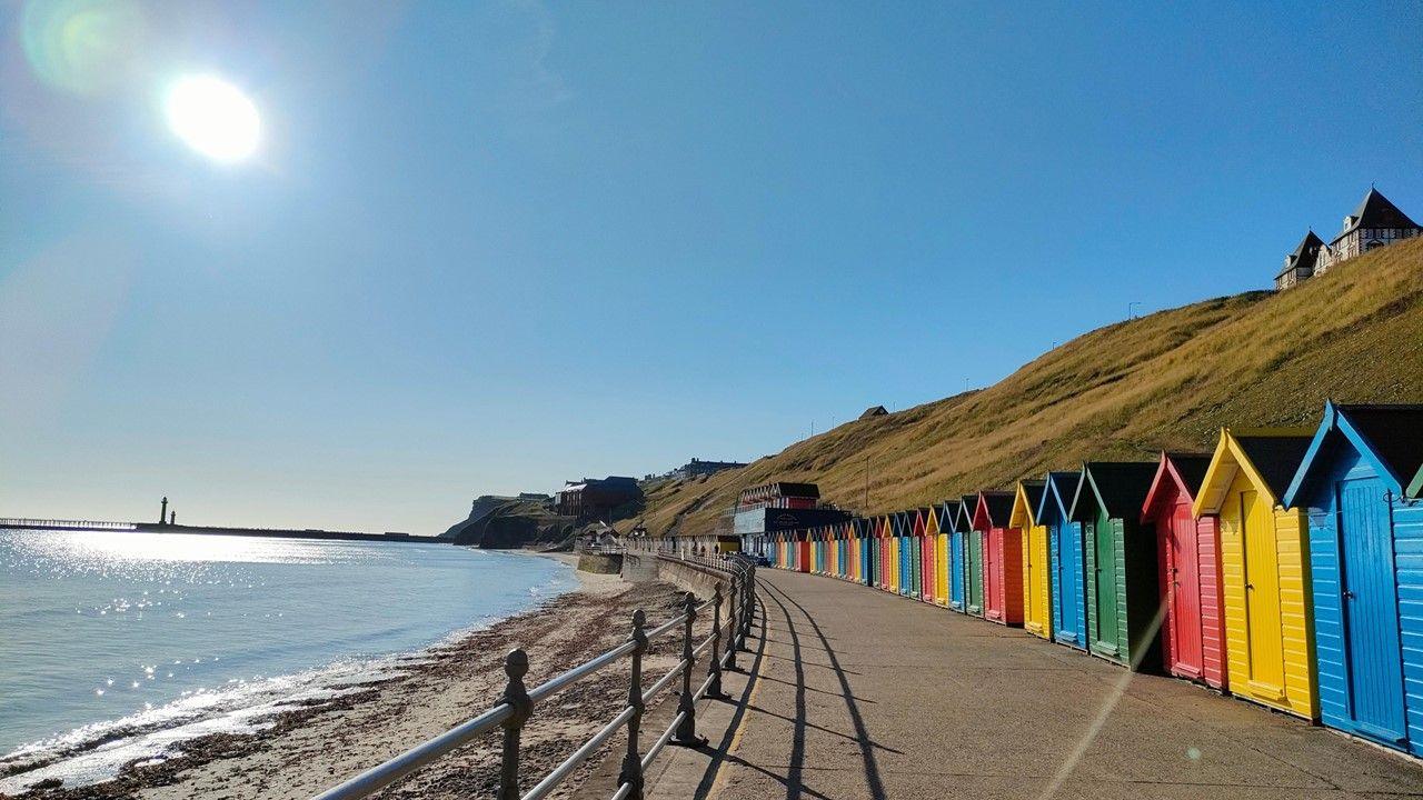 The Sun beats down from a blue sky over the sea and a row of coloured beach huts