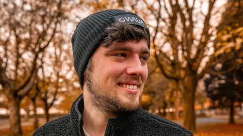 Cai Rhys Photograph of Cai Rhys, 26, taken in a autumnal park. Faded bare trees can be seen in the background, with orange leaves covering the floor. He wears a black beanie hat reading "Gwalia" and smiles away from the camera. He has brown eyes and a brown beard.