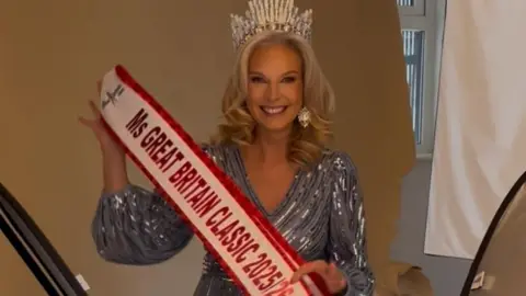 Laura White Laura behind the scenes of a photoshoot for Ms Great Britain Classic 2025. She is wearing a blue sparkly dress with a silver tiara and is holding up her sash.
