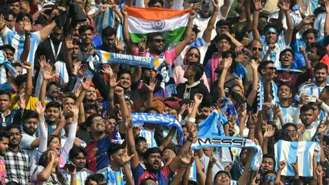Getty Images photo shows rows of people cheering and waving scarves at the Lionel Messi event in India.
